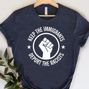 May include: Navy blue t-shirt with a white graphic. The graphic features a raised fist inside a circle with the text "KEEP THE IMMIGRANTS" above and "DEPORT THE RACISTS" below. Two stars flank the circle.