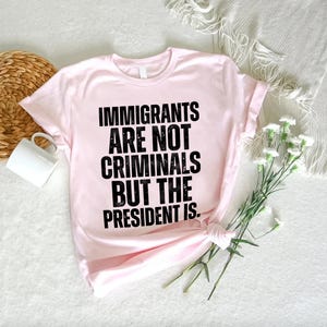 K&ouml;nnte beinhalten: Ein hellrosa T-Shirt mit dem Text "IMMIGRANTS ARE NOT CRIMINALS BUT THE PRESIDENT IS" in fetten schwarzen Buchstaben. Das T-Shirt ist am Saum geknotet. Eine wei&szlig;e Tasse und ein Weidenkorb sind im Hintergrund zu sehen.