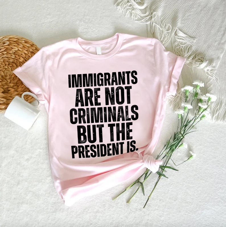 K&ouml;nnte beinhalten: Ein hellrosa T-Shirt mit dem Text "IMMIGRANTS ARE NOT CRIMINALS BUT THE PRESIDENT IS" in fetten schwarzen Buchstaben. Das T-Shirt ist am Saum geknotet. Eine wei&szlig;e Tasse und ein Weidenkorb sind im Hintergrund zu sehen.