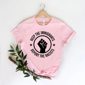 K&ouml;nnte beinhalten: Ein hellrosa T-Shirt mit dem Text "KEEP THE IMMIGRANTS, DEPORT THE RACISTS" in einem schwarzen Kreis-Design mit einer erhobenen Faust. Das T-Shirt h&auml;ngt an einem Holzkleiderb&uuml;gel.