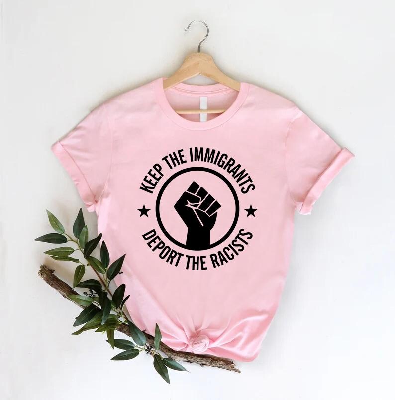 K&ouml;nnte beinhalten: Ein hellrosa T-Shirt mit dem Text "KEEP THE IMMIGRANTS, DEPORT THE RACISTS" in einem schwarzen Kreis-Design mit einer erhobenen Faust. Das T-Shirt h&auml;ngt an einem Holzkleiderb&uuml;gel.