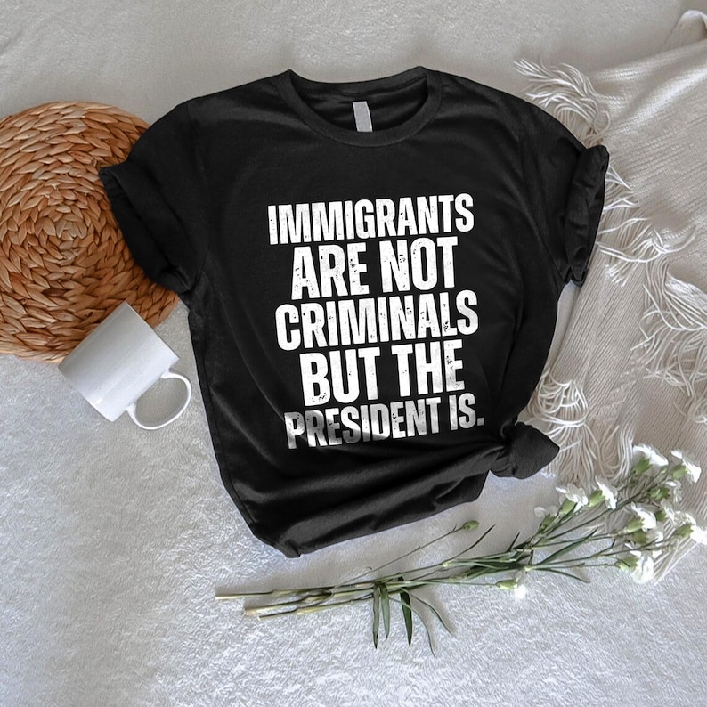 K&ouml;nnte beinhalten: Schwarzes T-Shirt mit wei&szlig;em Text: &bdquo;IMMIGRANTS ARE NOT CRIMINALS BUT THE PRESIDENT IS.&ldquo; Das T-Shirt ist gefaltet. Eine wei&szlig;e Tasse, eine gewebte Matte und Blumen sind im Hintergrund.