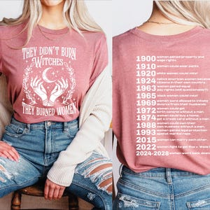 以下が含まれることがあります： 「THEY DIDN'T BURN WITCHES THEY BURNED WOMEN」の白文字がプリントされた、モーヴ色のTシャツ。デザインには三日月と手が含まれています。Tシャツの背面には、1900年から2028年までの女性の権利に関するマイルストーンが記載されています。