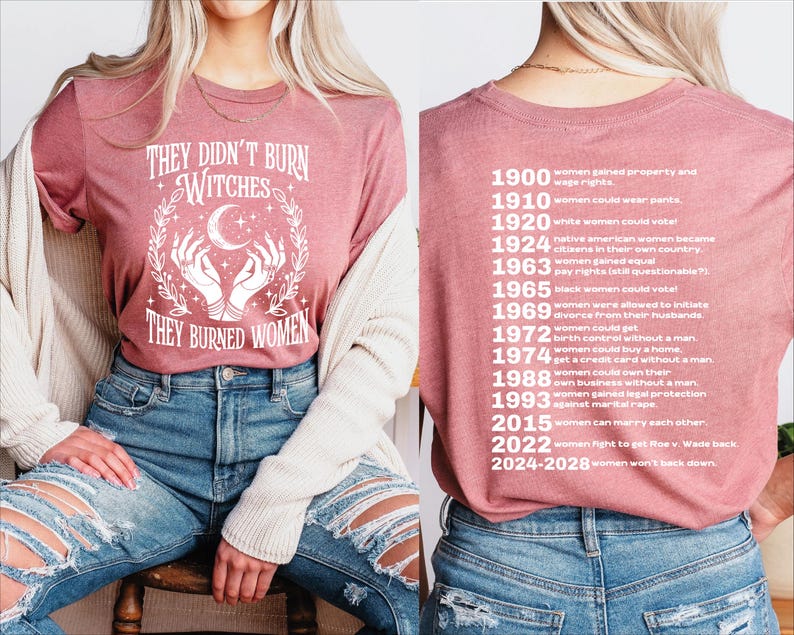 以下が含まれることがあります： 「THEY DIDN'T BURN WITCHES THEY BURNED WOMEN」の白文字がプリントされた、モーヴ色のTシャツ。デザインには三日月と手が含まれています。Tシャツの背面には、1900年から2028年までの女性の権利に関するマイルストーンが記載されています。