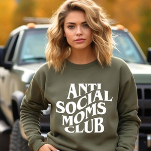 Puede incluir: Una mujer con una sudadera verde con el texto "Anti Social Moms Club" en blanco. Est&aacute; de pie frente a una camioneta.