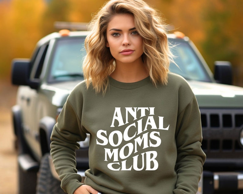 Puede incluir: Una mujer con una sudadera verde con el texto "Anti Social Moms Club" en blanco. Est&aacute; de pie frente a una camioneta.