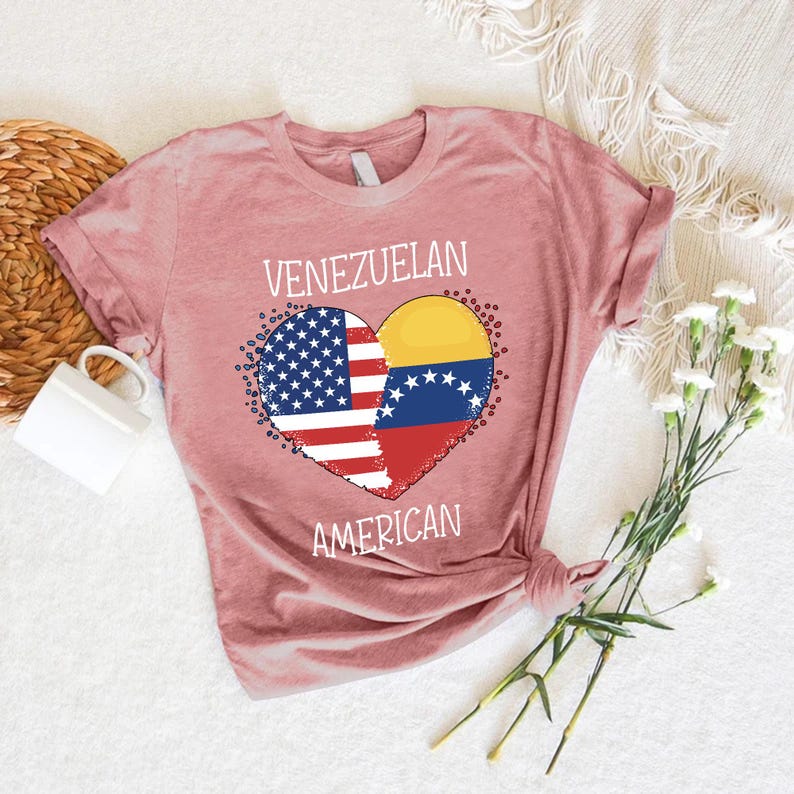 May include: A dusty rose t-shirt featuring a heart-shaped design with the flags of the United States and Venezuela. The words "VENEZUELAN" and "AMERICAN" are printed above and below the heart. The shirt is made of soft material.