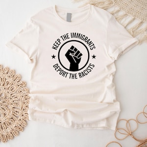 K&ouml;nnte beinhalten: Cremefarbenes T-Shirt mit schwarzem Aufdruck. Der Aufdruck zeigt eine geballte Faust in einem Kreis, mit den Worten "KEEP THE IMMIGRANTS" dar&uuml;ber und "DEPORT THE RACISTS" darunter. Zwei kleine Sterne flankieren die Faust.