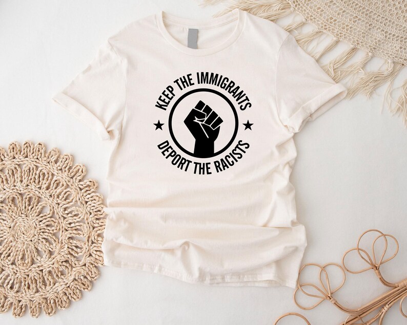 K&ouml;nnte beinhalten: Cremefarbenes T-Shirt mit schwarzem Aufdruck. Der Aufdruck zeigt eine geballte Faust in einem Kreis, mit den Worten "KEEP THE IMMIGRANTS" dar&uuml;ber und "DEPORT THE RACISTS" darunter. Zwei kleine Sterne flankieren die Faust.