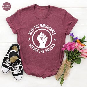 K&ouml;nnte beinhalten: Weinrotes T-Shirt mit dem Text "KEEP THE IMMIGRANTS, DEPORT THE RACISTS" in einem kreisf&ouml;rmigen Design mit einer erhobenen Faust. Schwarz-wei&szlig;e Turnschuhe liegen neben dem Shirt. Ein Blumenstrau&szlig; ist ebenfalls im Bild.
