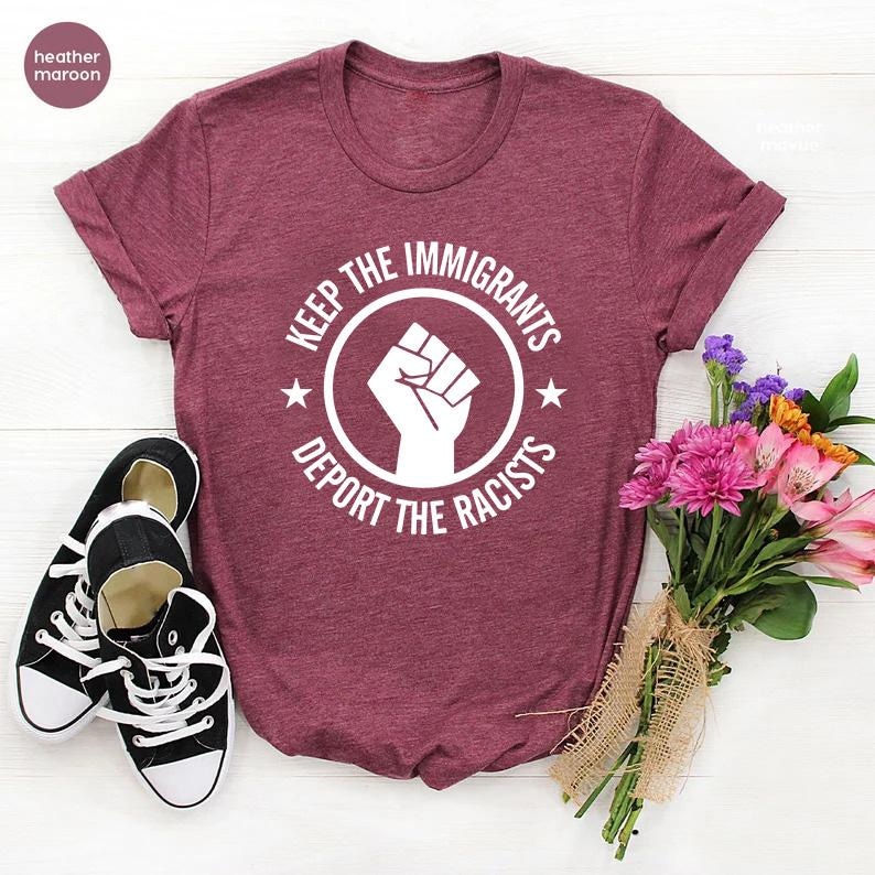 K&ouml;nnte beinhalten: Weinrotes T-Shirt mit dem Text "KEEP THE IMMIGRANTS, DEPORT THE RACISTS" in einem kreisf&ouml;rmigen Design mit einer erhobenen Faust. Schwarz-wei&szlig;e Turnschuhe liegen neben dem Shirt. Ein Blumenstrau&szlig; ist ebenfalls im Bild.