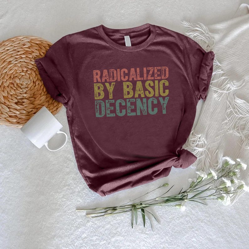 Puede incluir: Camiseta burdeos con el texto "RADICALIZED BY BASIC DECENCY" en una fuente retro. La camiseta tiene las mangas enrolladas y un nudo en la parte inferior. Una taza blanca, una cesta de mimbre y flores est&aacute;n en el fondo.