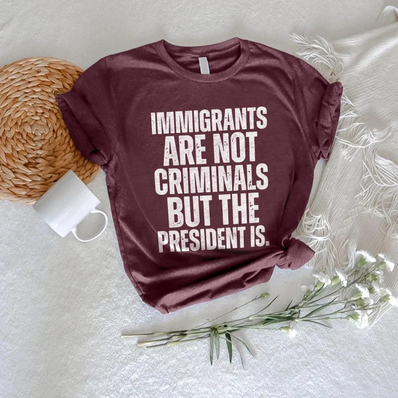 K&ouml;nnte beinhalten: Burgunderfarbenes T-Shirt mit dem Text "IMMIGRANTS ARE NOT CRIMINALS BUT THE PRESIDENT IS" in wei&szlig;en Blockbuchstaben. Das Shirt ist gefaltet und liegt auf einer wei&szlig;en Oberfl&auml;che mit einem gewebten Untersetzer, einer wei&szlig;en Tasse und einem Zweig wei&szlig;er Blumen.