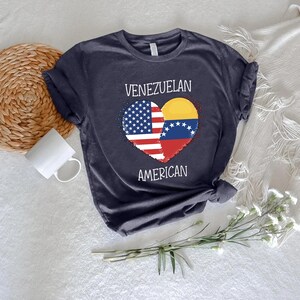 May include: Navy blue t-shirt featuring a heart-shaped design split between the American and Venezuelan flags. The words "VENEZUELAN" and "AMERICAN" are printed above and below the heart. The shirt is made of soft material.