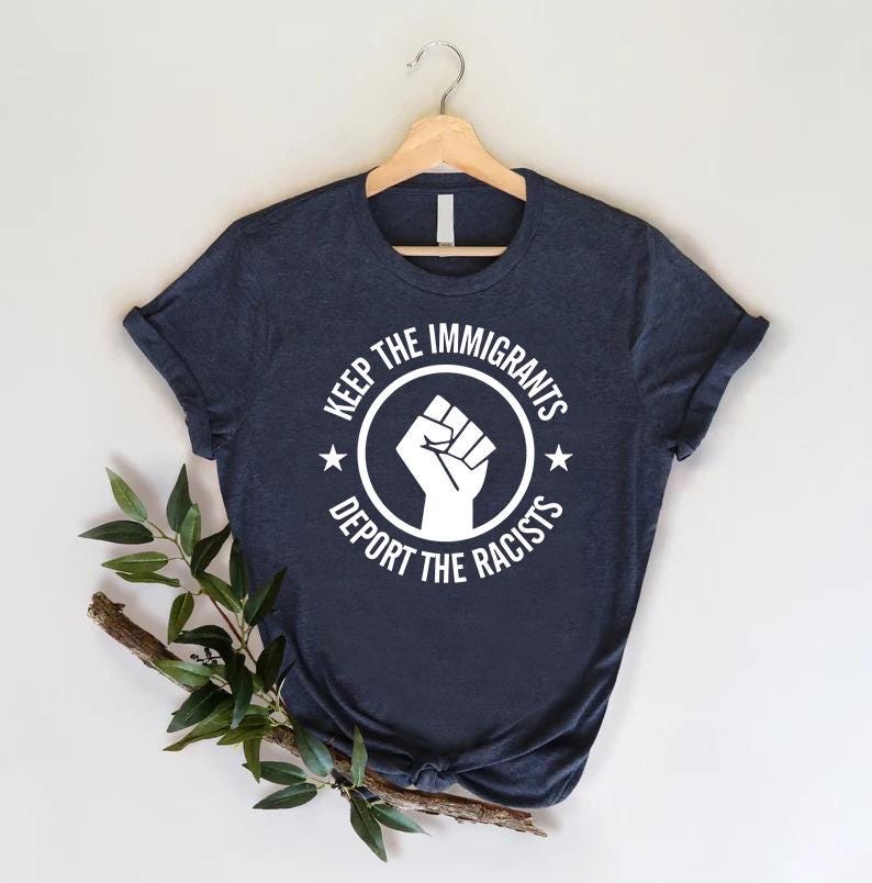 K&ouml;nnte beinhalten: Marineblaues T-Shirt mit wei&szlig;em Aufdruck. Der Aufdruck zeigt eine geballte Faust in einem Kreis mit dem Text "KEEP THE IMMIGRANTS" dar&uuml;ber und "DEPORT THE RACISTS" darunter. Zwei Sterne flankieren den Kreis.