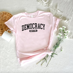 May include: A light pink t-shirt with the word "DEMOCRACY" printed in bold black letters. Below the word "DEMOCRACY" is the text "EST 1776" in a black rectangle. The shirt is folded and tied at the bottom.