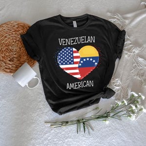May include: Black t-shirt featuring a heart-shaped design with the American and Venezuelan flags. The words "VENEZUELAN" and "AMERICAN" are printed above and below the heart in white. The shirt is displayed on a white surface.