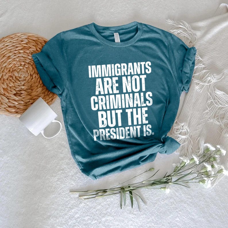 K&ouml;nnte beinhalten: T&uuml;rkisfarbenes T-Shirt mit dem wei&szlig;en Text "IMMIGRANTS ARE NOT CRIMINALS BUT THE PRESIDENT IS". Das Shirt hat hochgekrempelte &Auml;rmel und einen Knoten am Saum. Eine wei&szlig;e Tasse und ein gewebter Untersetzer sind im Hintergrund zu sehen.