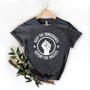 K&ouml;nnte beinhalten: Dunkelgraues T-Shirt mit wei&szlig;er Grafik. Die Grafik zeigt eine geballte Faust in einem Kreis mit dem Text "KEEP THE IMMIGRANTS" dar&uuml;ber und "DEPORT THE RACISTS" darunter. Zwei Sterne flankieren die Faust.