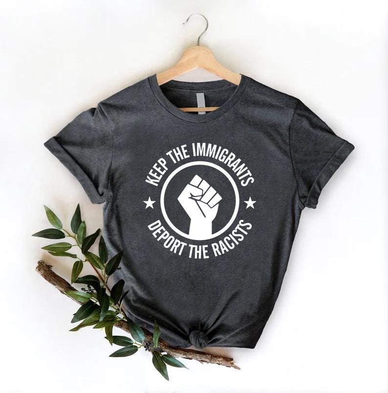 K&ouml;nnte beinhalten: Dunkelgraues T-Shirt mit wei&szlig;er Grafik. Die Grafik zeigt eine geballte Faust in einem Kreis mit dem Text "KEEP THE IMMIGRANTS" dar&uuml;ber und "DEPORT THE RACISTS" darunter. Zwei Sterne flankieren die Faust.
