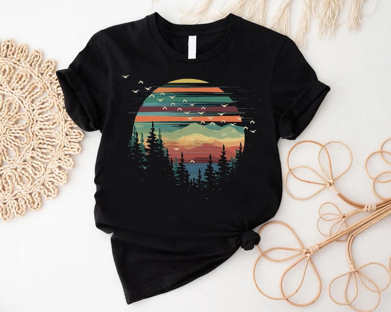 May include: Black t-shirt featuring a colorful graphic of a sunset over a mountain range with pine trees. The design includes a vibrant sky with birds in flight, creating a nature-inspired scene.