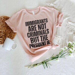 K&ouml;nnte beinhalten: Hellrosa T-Shirt mit dem schwarzen Text: &bdquo;IMMIGRANTS ARE NOT CRIMINALS BUT THE PRESIDENT IS.&ldquo; Das Shirt liegt auf einer wei&szlig;en Oberfl&auml;che, zusammen mit einer wei&szlig;en Tasse, einem geflochtenen Korb und Blumen.