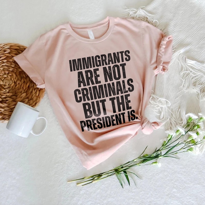 K&ouml;nnte beinhalten: Hellrosa T-Shirt mit dem schwarzen Text: &bdquo;IMMIGRANTS ARE NOT CRIMINALS BUT THE PRESIDENT IS.&ldquo; Das Shirt liegt auf einer wei&szlig;en Oberfl&auml;che, zusammen mit einer wei&szlig;en Tasse, einem geflochtenen Korb und Blumen.