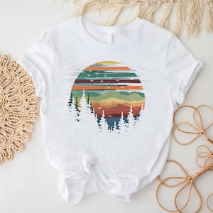 May include: White t-shirt featuring a colorful graphic of a sunset over a forest. The design includes horizontal stripes in shades of orange, green, and blue, with silhouettes of trees and flying birds. The shirt is made of a soft material.