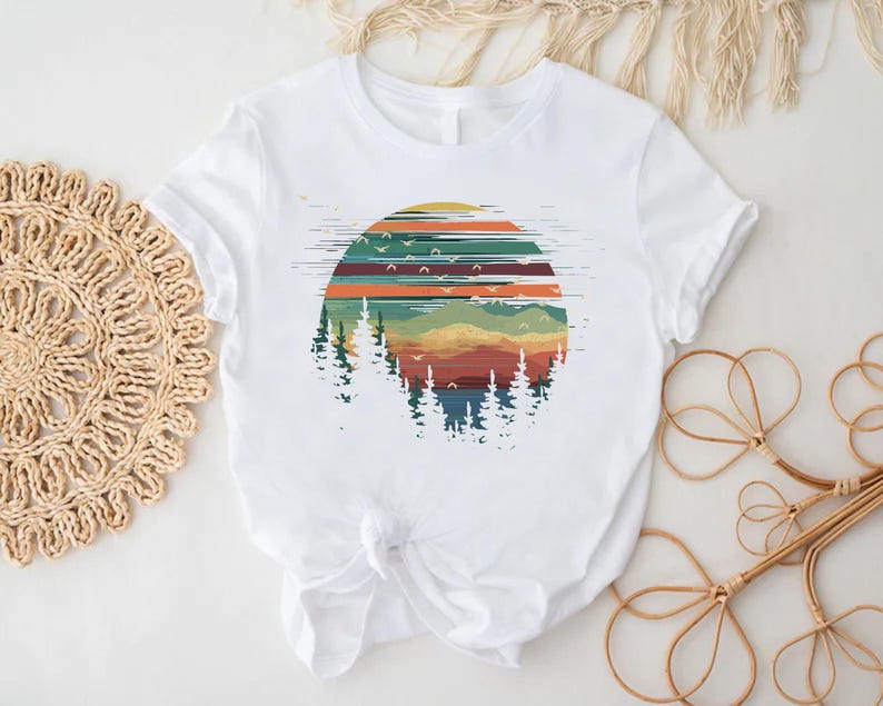 May include: White t-shirt featuring a colorful graphic of a sunset over a forest. The design includes horizontal stripes in shades of orange, green, and blue, with silhouettes of trees and flying birds. The shirt is made of a soft material.