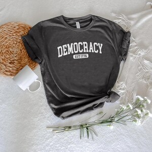 May include: Dark gray t-shirt with the word "DEMOCRACY" in white, arched text. Below the word "DEMOCRACY" is the text "EST 1776". The shirt is knotted at the bottom and is on a white surface.