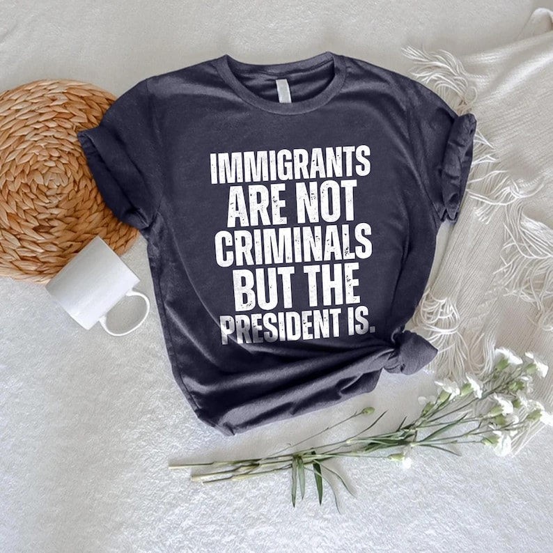 K&ouml;nnte beinhalten: Dunkelblaues T-Shirt mit dem wei&szlig;en Text: "IMMIGRANTS ARE NOT CRIMINALS BUT THE PRESIDENT IS." Das Shirt wird auf einer wei&szlig;en Oberfl&auml;che mit einem gewebten Untersetzer, einer wei&szlig;en Tasse und einem Zweig wei&szlig;er Blumen pr&auml;sentiert.