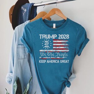 May include: Teal-colored t-shirt with a distressed American flag design. The shirt features the text "TRUMP 2028," "We The People," and "KEEP AMERICA GREAT." The design includes the numbers 45, 47, and 48.