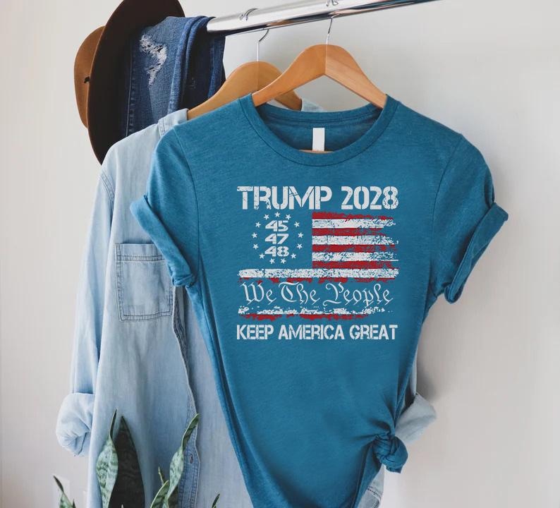May include: Teal-colored t-shirt with a distressed American flag design. The shirt features the text "TRUMP 2028," "We The People," and "KEEP AMERICA GREAT." The design includes the numbers 45, 47, and 48.
