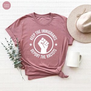 K&ouml;nnte beinhalten: Staubrosa T-Shirt mit wei&szlig;er Grafik eines erhobenen F&auml;ustlings in einem Kreis, mit dem Text "KEEP THE IMMIGRANTS" dar&uuml;ber und "DEPORT THE RACISTS" darunter. Zwei Sterne flankieren die Faust. Das Shirt ist gefaltet.