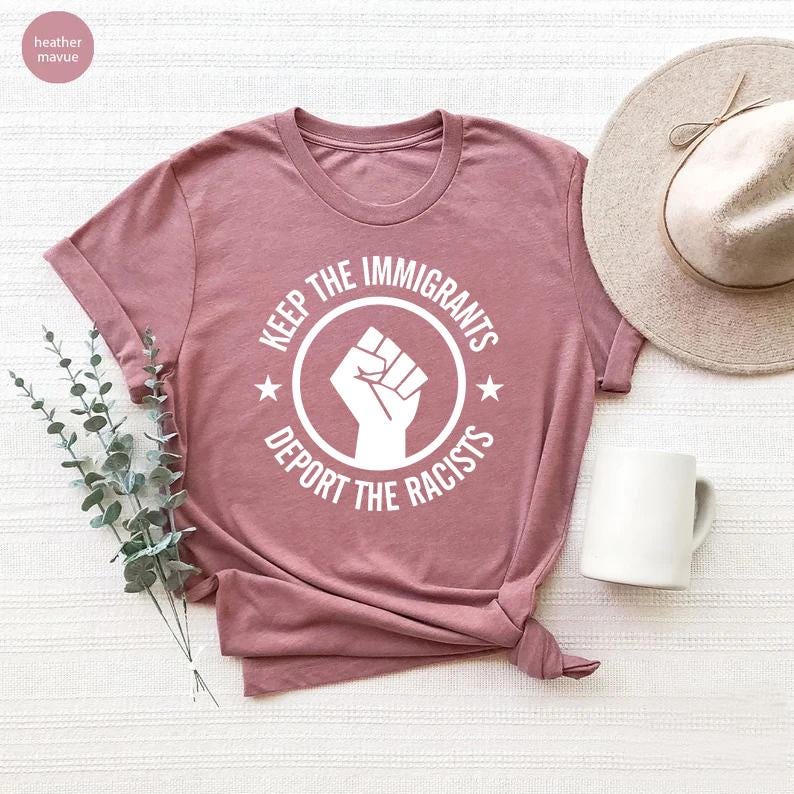 K&ouml;nnte beinhalten: Staubrosa T-Shirt mit wei&szlig;er Grafik eines erhobenen F&auml;ustlings in einem Kreis, mit dem Text "KEEP THE IMMIGRANTS" dar&uuml;ber und "DEPORT THE RACISTS" darunter. Zwei Sterne flankieren die Faust. Das Shirt ist gefaltet.