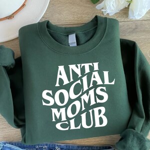Puede incluir: Una sudadera verde oscuro con texto blanco que dice "Anti Social Moms Club" en una fuente retro.