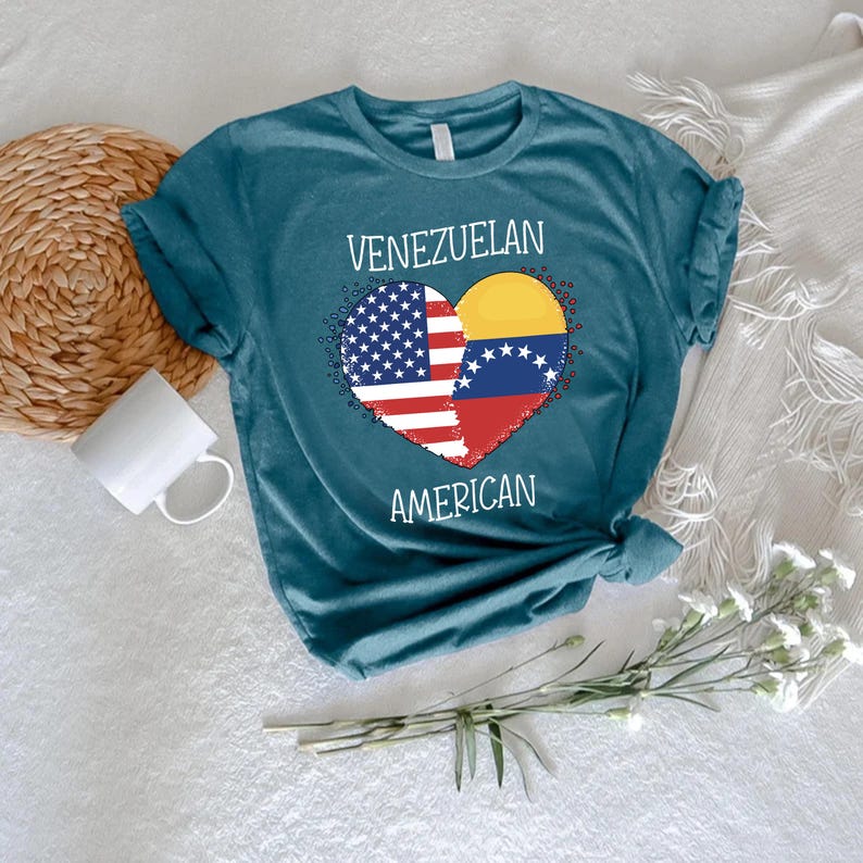 May include: Teal t-shirt featuring a heart design split between the American and Venezuelan flags. The words "VENEZUELAN" and "AMERICAN" are arched above and below the heart. The shirt is displayed on a white surface with a woven basket and a white mug.
