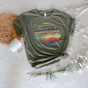 May include: Olive green t-shirt featuring a colorful sunset design with mountains, trees, and flying birds. The graphic has horizontal stripes of orange, red, and teal. The shirt is knotted at the side.