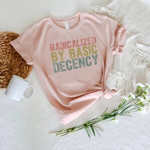 Puede incluir: Una camiseta rosa claro con el texto "RADICALIZED BY BASIC DECENCY" en una fuente retro. La camiseta est&aacute; anudada en la parte inferior y se muestra sobre una superficie blanca con una cesta tejida, una taza blanca y flores.