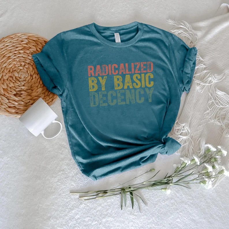Puede incluir: Camiseta de color turquesa con el texto "RADICALIZED BY BASIC DECENCY" en rojo, amarillo y verde. La camiseta est&aacute; anudada en la parte inferior y tiene las mangas enrolladas. Una taza blanca y un posavasos tejido est&aacute;n en el fondo.