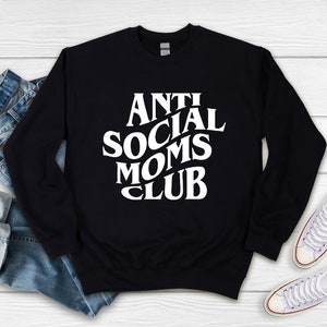 Puede incluir: Sudadera negra con texto blanco que dice "Anti Social Moms Club". La sudadera est&aacute; colocada sobre una superficie de madera blanca con un par de zapatillas blancas y unos vaqueros azules.