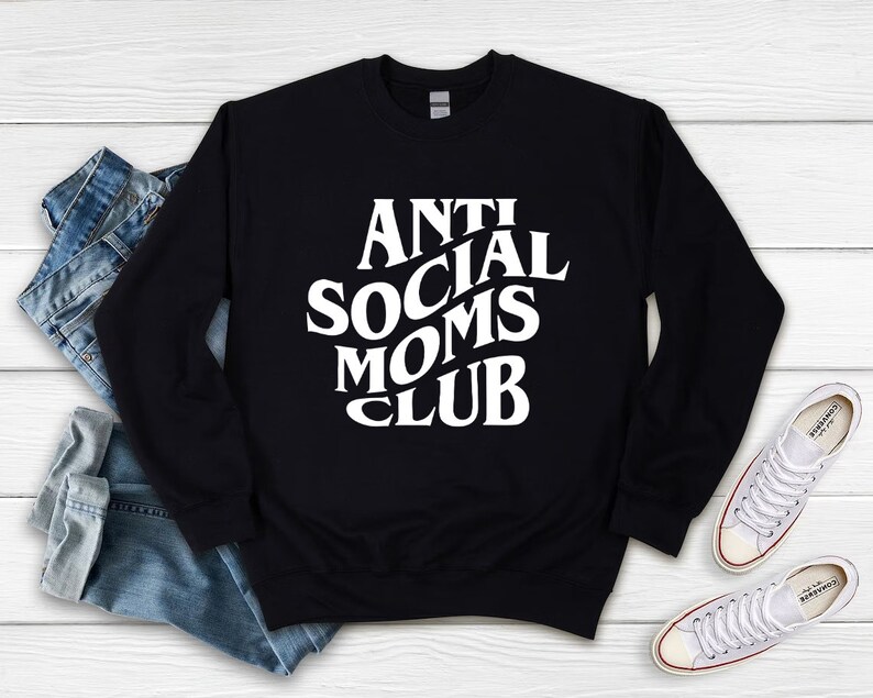 Puede incluir: Sudadera negra con texto blanco que dice "Anti Social Moms Club". La sudadera est&aacute; colocada sobre una superficie de madera blanca con un par de zapatillas blancas y unos vaqueros azules.
