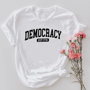 May include: White t-shirt with the word "DEMOCRACY" in bold black letters, with "EST 1776" below it. The shirt is styled with rolled-up sleeves and a knot at the bottom. Pink flowers are arranged to the right of the shirt.