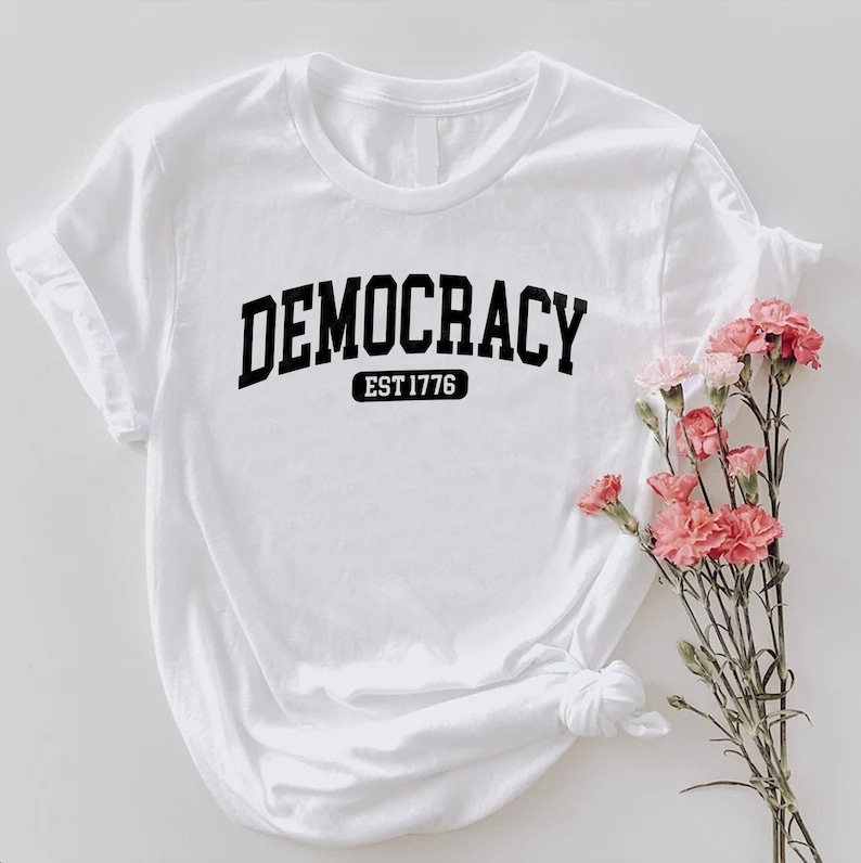 May include: White t-shirt with the word "DEMOCRACY" in bold black letters, with "EST 1776" below it. The shirt is styled with rolled-up sleeves and a knot at the bottom. Pink flowers are arranged to the right of the shirt.
