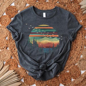 May include: Dark gray t-shirt featuring a colorful sunset design with mountains, trees, and flying birds. The graphic includes shades of orange, yellow, green, and blue. The shirt is knotted at the waist and is displayed on a woven, circular rug.