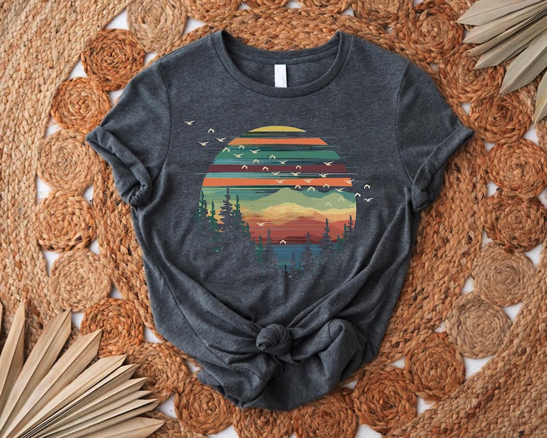 May include: Dark gray t-shirt featuring a colorful sunset design with mountains, trees, and flying birds. The graphic includes shades of orange, yellow, green, and blue. The shirt is knotted at the waist and is displayed on a woven, circular rug.