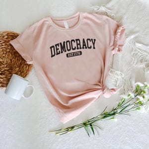 May include: A light pink t-shirt with the word "DEMOCRACY" printed in black arched letters. Below the word "DEMOCRACY" is the text "EST 1776" in a black rectangular box. The shirt is knotted at the bottom.