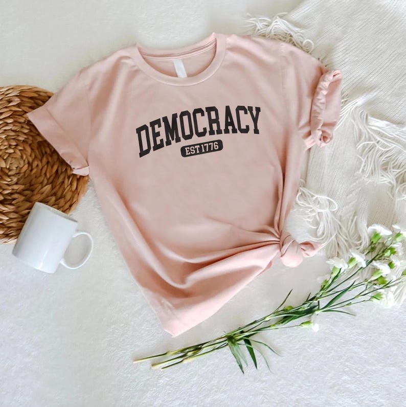 May include: A light pink t-shirt with the word "DEMOCRACY" printed in black arched letters. Below the word "DEMOCRACY" is the text "EST 1776" in a black rectangular box. The shirt is knotted at the bottom.