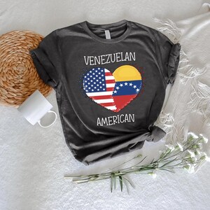 May include: Dark gray t-shirt featuring a heart design split between the American and Venezuelan flags. The words "VENEZUELAN" and "AMERICAN" are printed above and below the heart. The shirt is made of soft material.