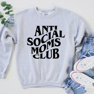 Puede incluir: Sudadera gris con texto negro que dice "Anti Social Moms Club" en una fuente retro. La sudadera est&aacute; colocada sobre una superficie blanca con un par de pantalones vaqueros azules y zapatillas blancas.